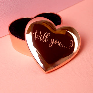 Rose gold engagement heart ring box with "Will you...?" engraved on the front, shown in close up shot.