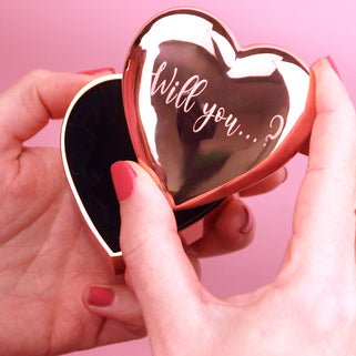 Rose gold engagement heart ring box with "Will you...?" engraved on the front, shown in close up shot with model. 