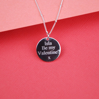 Silver Circle Disc Necklace with personalised "Isla Be my Valentine? x" text engraving, on a satellite chain shown in a close up shot