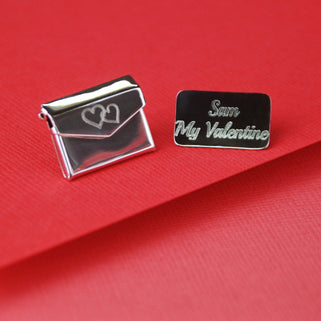 Love letter secret message pin with a double heart icon engraved on the front of the envelope and "Sam My Valentine" engraved on the letter, shown in close up shot on red background.