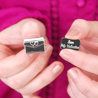 Love letter secret message pin with a double heart icon engraved on the front of the envelope and "Sam My Valentine" engraved on the letter, shown in close up with model.