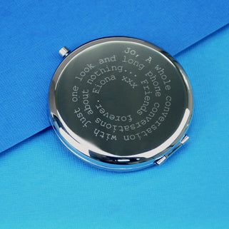 Silver compact mirror with personalised spiral message in typewriter font engraved on the front, shown in close up shot on blue background.