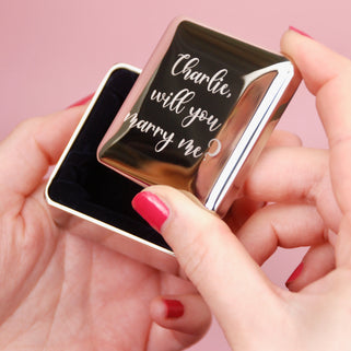 Silver ring box with personalised message "Charlie, will you marry me?" engraved on the front, shown in close up shot with model