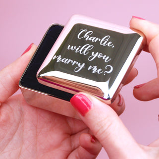 Silver ring box with personalised message "Charlie, will you marry me?" engraved on the front, shown in close up shot with model